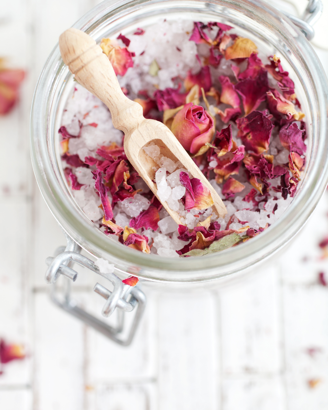 Top-down view of a glass clamp-lid jar filled with white bath salts mixed with dried pink and red rose petals, with a small wooden scoop resting inside.