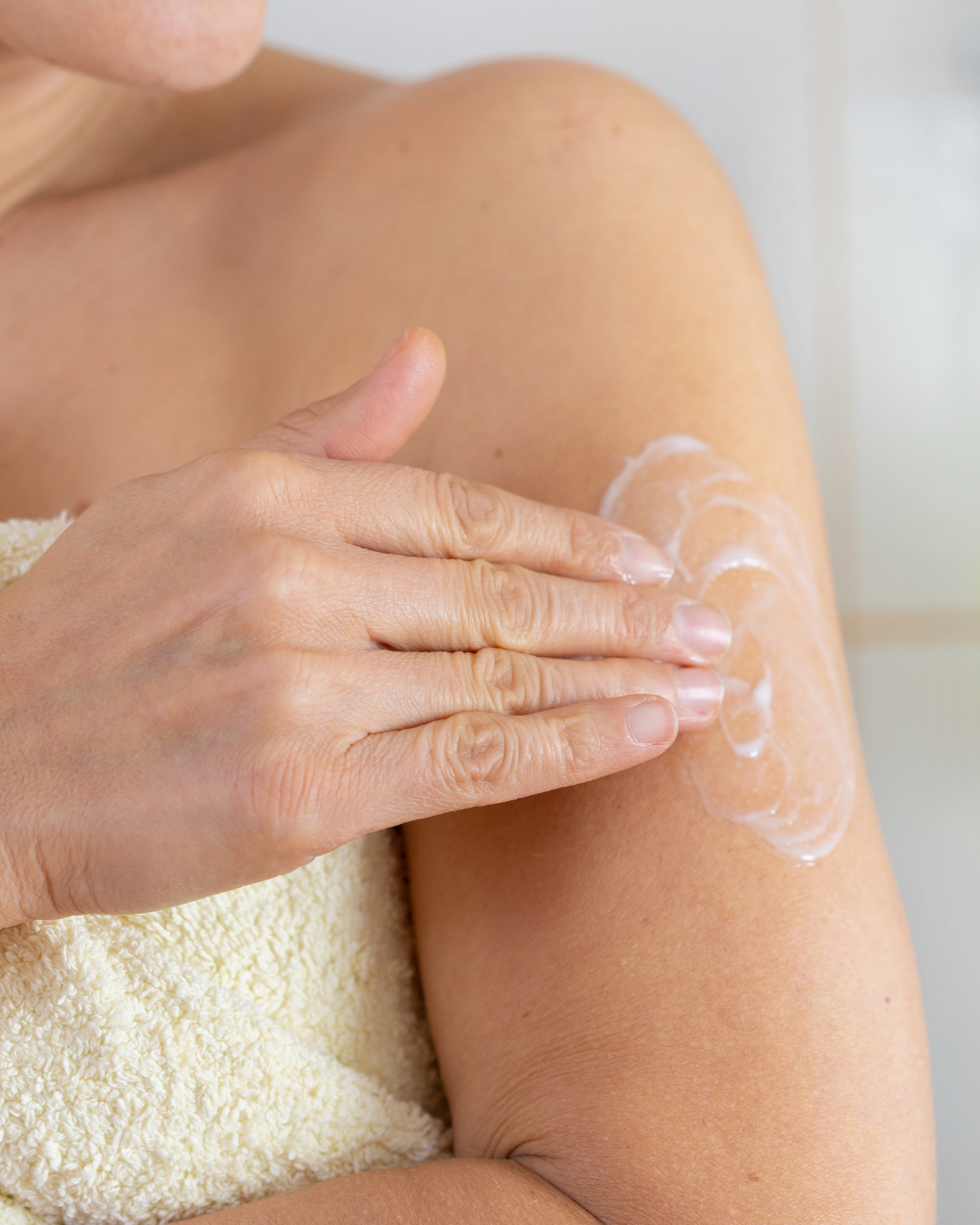 A close-up of a person applying white lotion or sunscreen cream onto their upper arm with one hand, wrapped in a white towel.