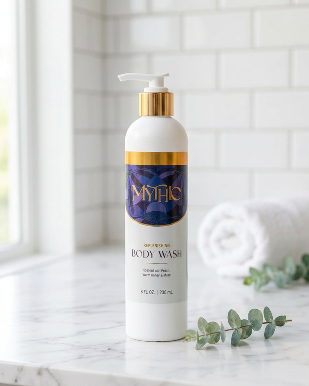 A bottle of Mythic Replenishing Body Wash standing on a marble countertop with a rolled towel and eucalyptus leaves in a bright bathroom setting.