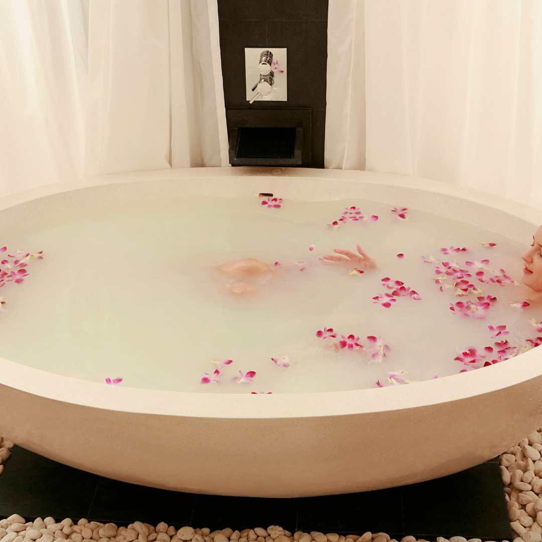 Bath tub with water and pink flower petals, surrounded by pebbles and a curtain showing woman using mythic's rose botanical soak